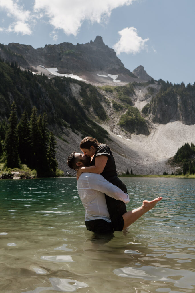 Groom lifting future bride and kissing her standing in waist deep water in an alpine lake with mountains in the background at Mount Rainier National Park.