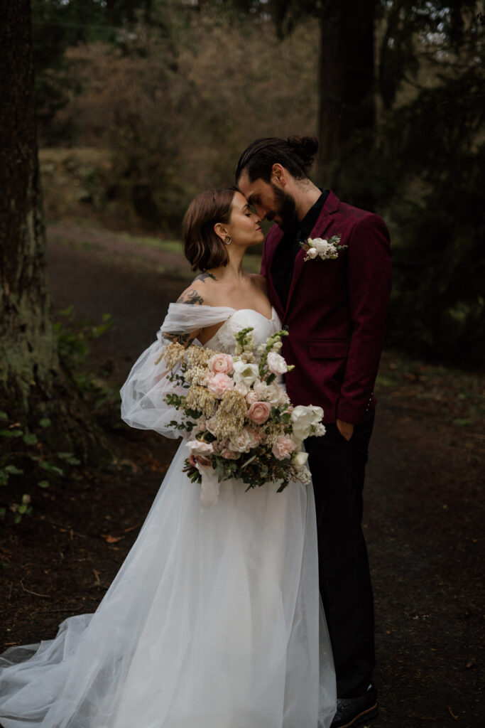 Couple standing forehead to forehead with huge bridal bouquet from centerpiece floral posed in their wedding attire during their elopement at Portland's Hoyt Arboretum.
