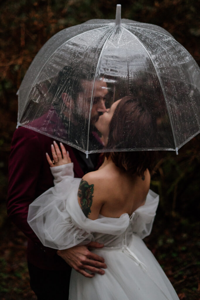 Bridal couple kissing on their elopement day under a clear umbrella in the rain during their Oregon Elopement.
