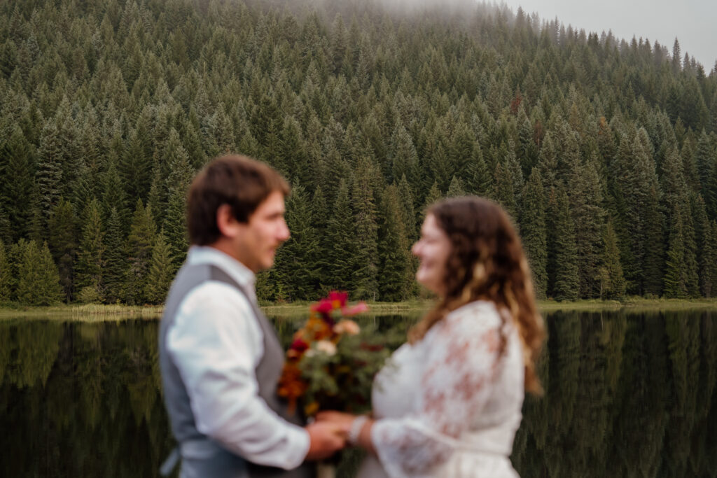 Foggy forest scenery in focus in the background and Bride and Groom out of focus holding hands and saying vows on the dock at Trillium Lake in Oregon after their sunrise canoe session.