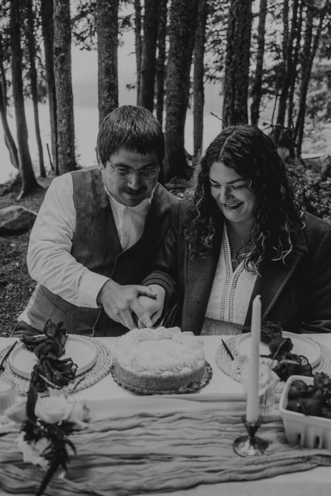 B&W photo of bride and groom cutting their elopement cake at a picnic table with Mount Hood National Forest and Trillium Lake in the background during their Canoe Elopement day.