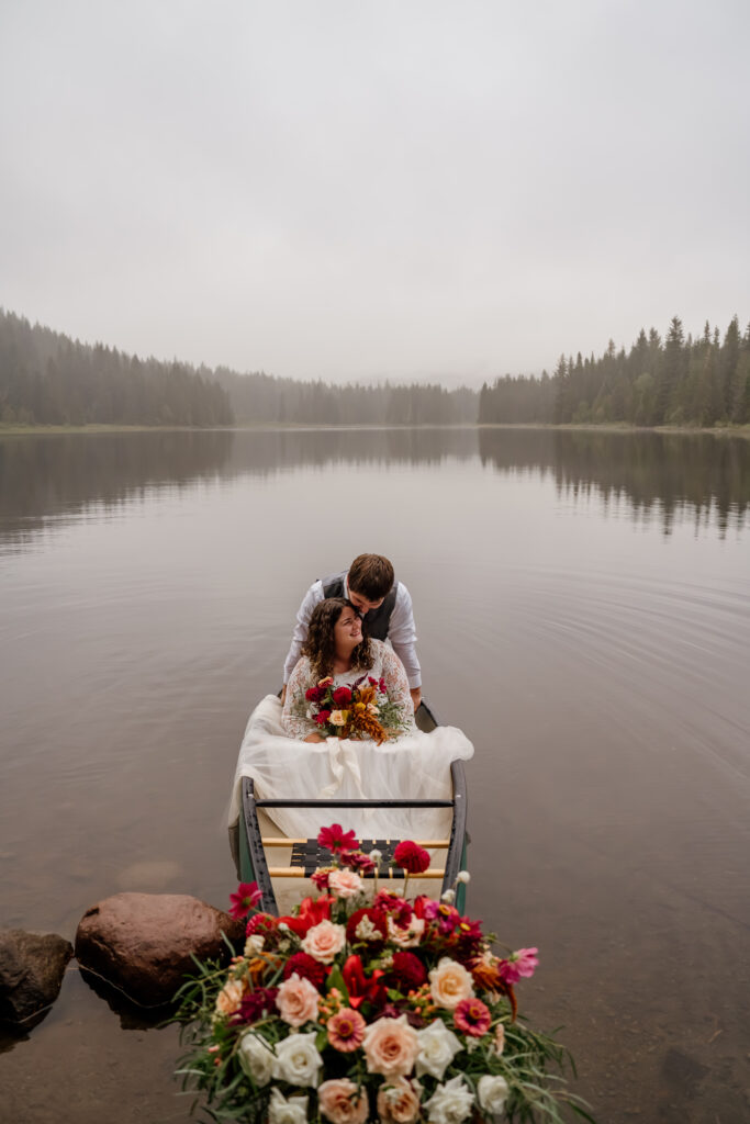 Groom hugging bride from behind while sitting in a canoe with a floral install during their sunrise canoe elopement at Trillium Lake in Oregon