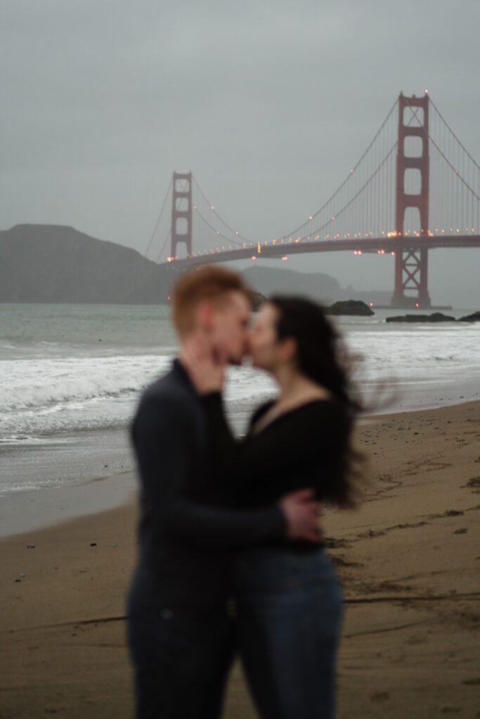 Couple in foreground and blurry with the Golden Gate Bridge in the background in focus during their engagement photos at Baker Beach.