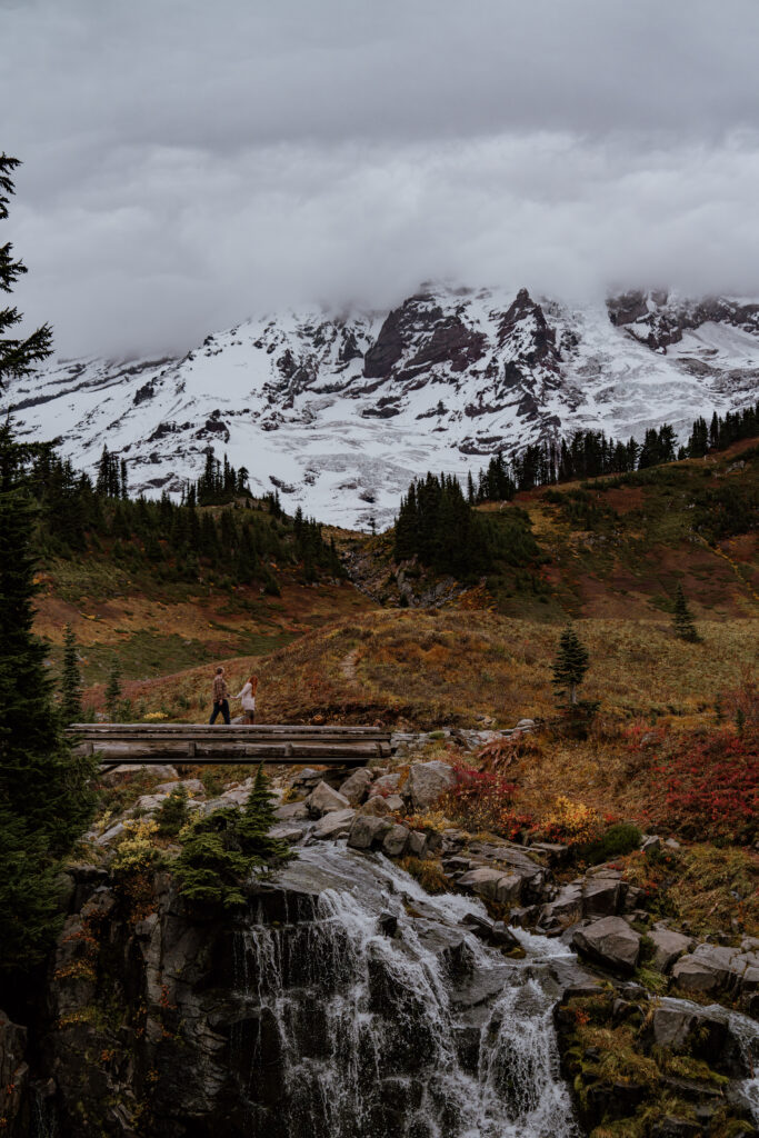 Couple walking across bridge with Mount Rainier towering in the background and surrounded by fall colors at Mount Rainier National Park.