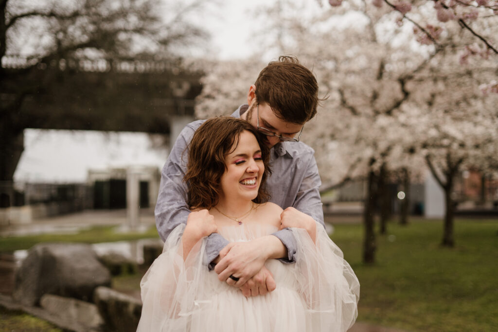 Couple standing snuggled in at Tom McCall Park during the Cherry Blossom bloom.