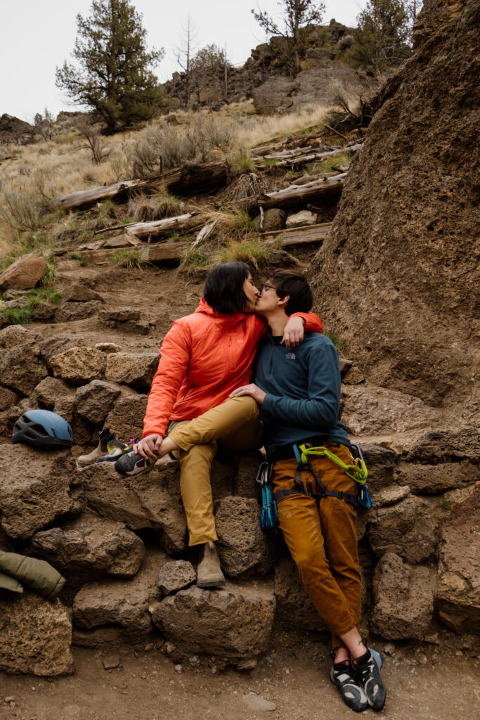 Couple sitting side by side and kissing during their climbing adventure session at smith rock state park.