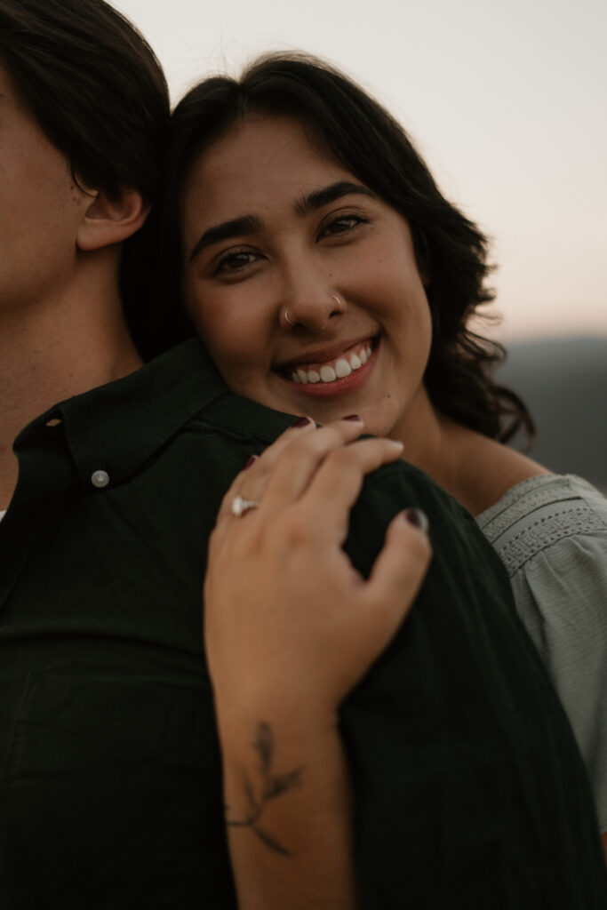 Womans face looking at camera while hugging her mans back during their Columbia River Gorge engagement photos.