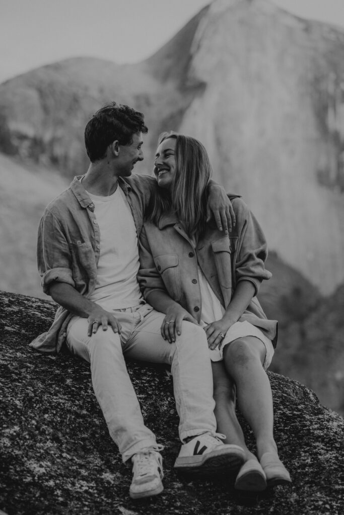 B+W photo of couple sitting and looking at each other smiling with Half Dome in the background.