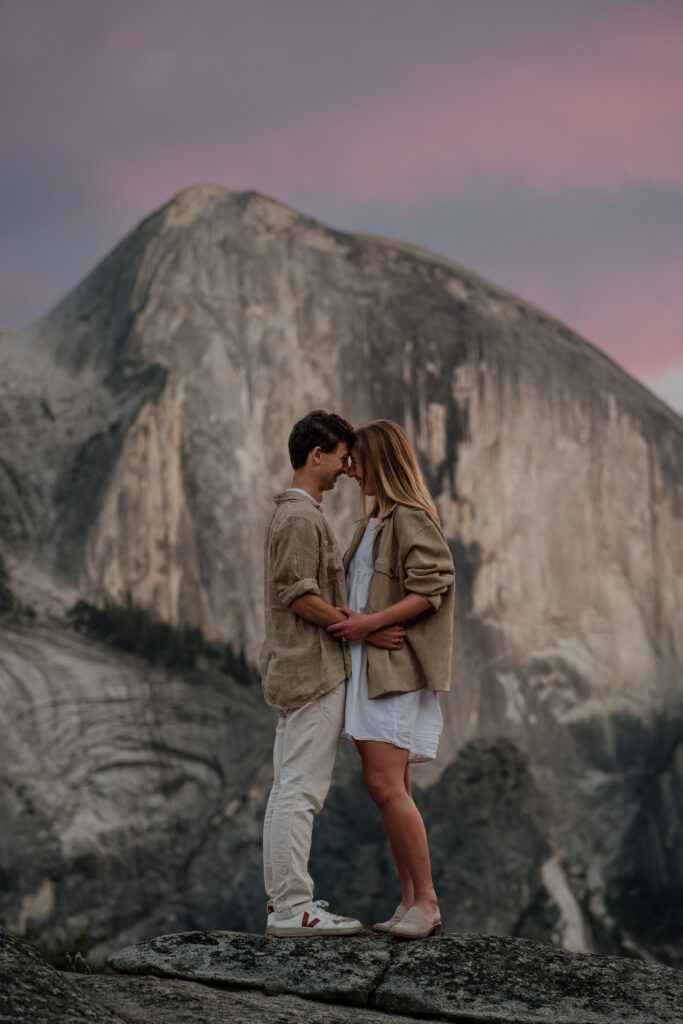 Couple standing forehead to forehead with Half dome in the background and a vibrant purple and pink sunset overhead.