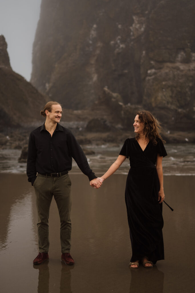 Couple standing and holding hands with Haystack rock in Cannon Beach in the background during their engagement session.