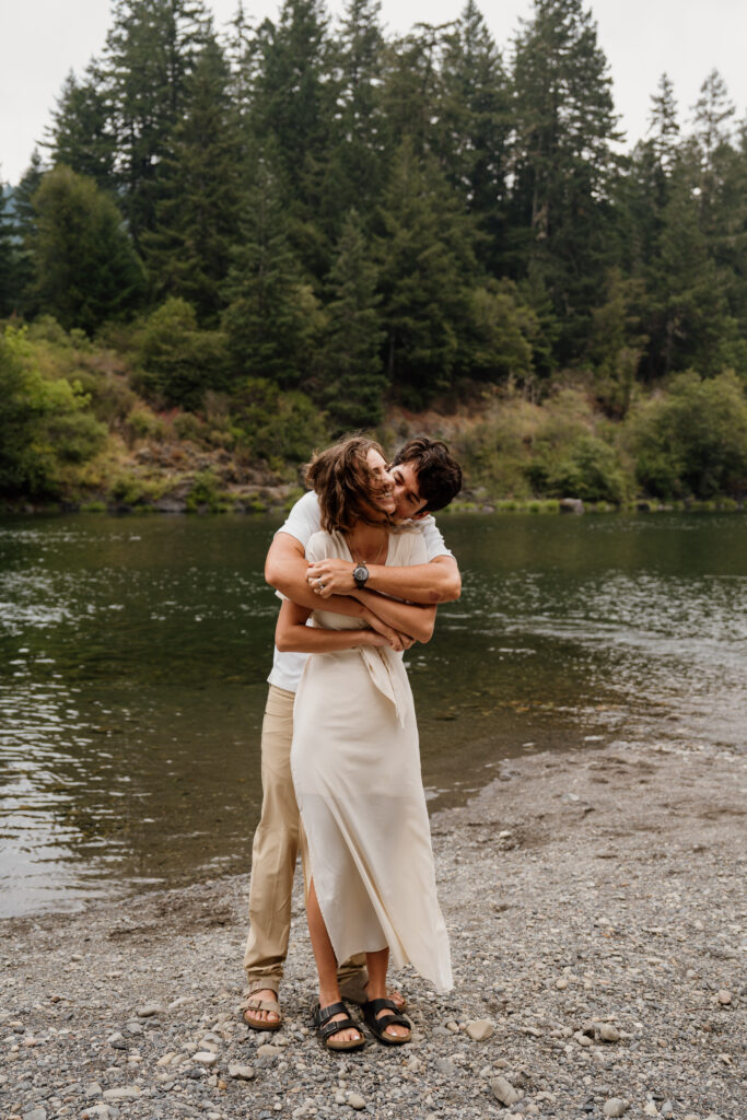 Groom hugging bride fro behind with a river in the background in Jedidiah Smith State Park in the redwoods during their hiking elopement.