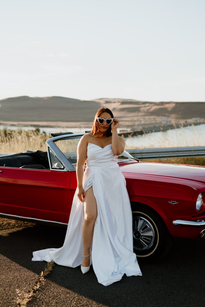 Woman in bridal attire posed in front of a classic cherry red ford mustang with the Columbia River Gorge in the background.