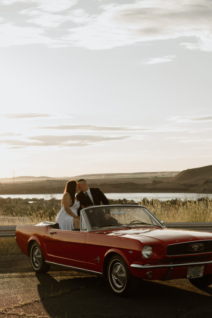 Couple sitting in the back of a Cherry Red Vintage Ford Mustang and kissing with the Columbia River Gorge in the background during sunset.