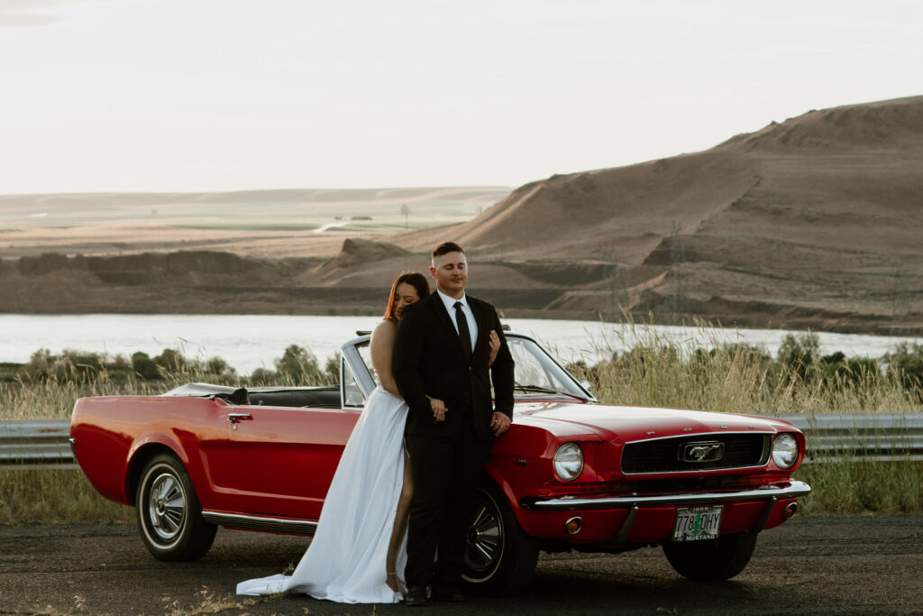 Woman in white dress leaning her head on the back of her partner with her eyes closed during with vintage red mustang and the Columbia River Gorge in the background.