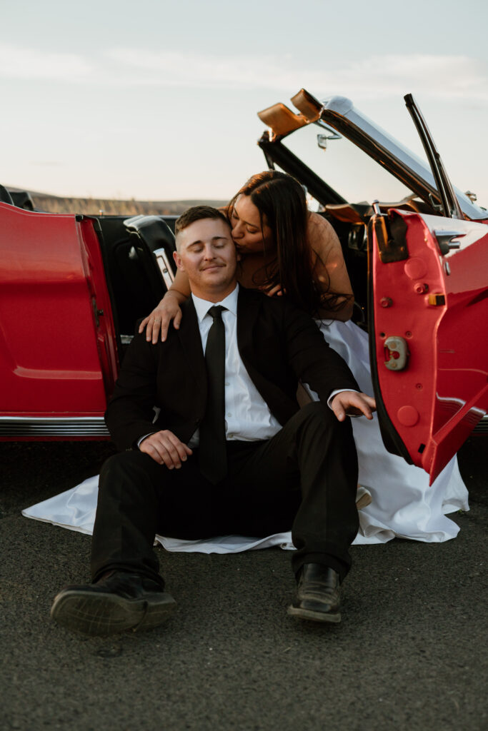 Man sitting on ground in front of woman with car door open getting a kiss on the temple from her during their Columbia River Gorge Vintage Car Engagement Photos.