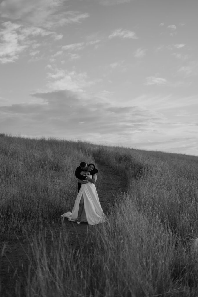 Black and white photo of man hugging woman partner from behind surrounded by beautiful tall grass during their engagement photos in the Columbia River Gorge.