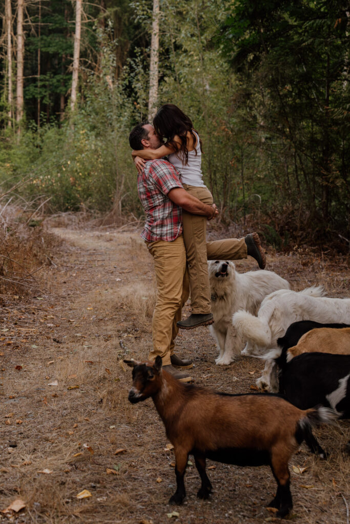 Man picking up woman in farm clothes and kissing her surrounded by two dogs and four goats.