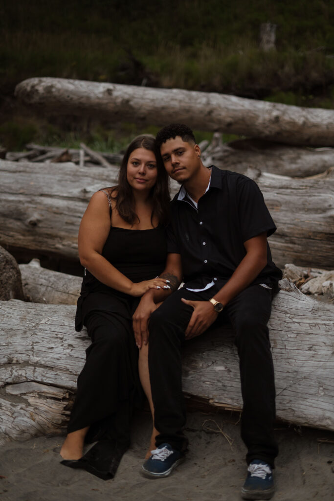 Couple sitting on driftwood logs on the beach with their heads leaned into each other while softly smiling for the camera.