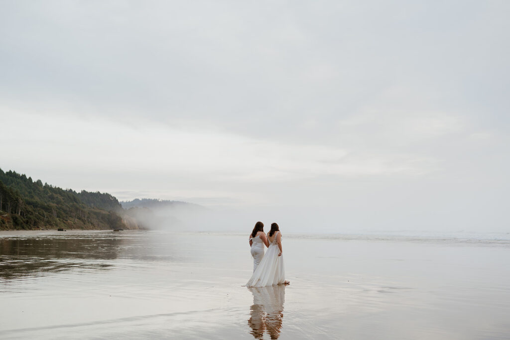 Two brides walk into the high tide with a foggy backdrop