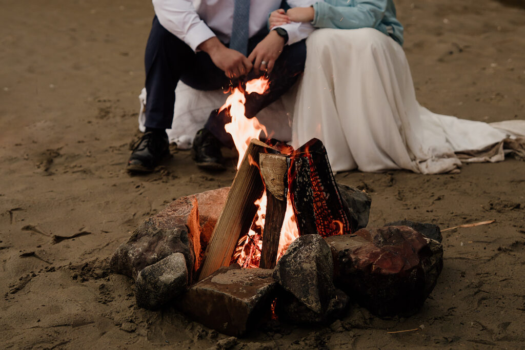 Bride and groom cuddle up by the fire on Arcadia Beach