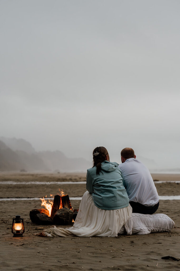 Bride and groom cuddle up by the fire on Arcadia Beach