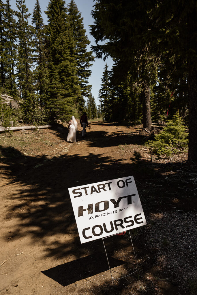 Bride and groom walk in front of a sign that reads "start of hoyt archery course"