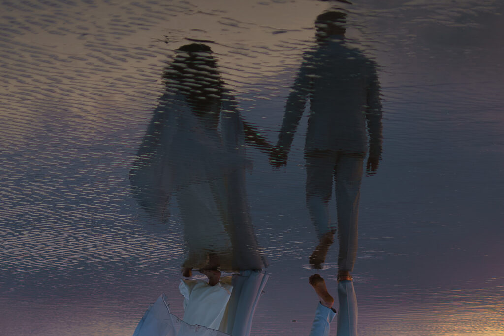 A reflection of bride and groom walking on the beach shows in the ocean water