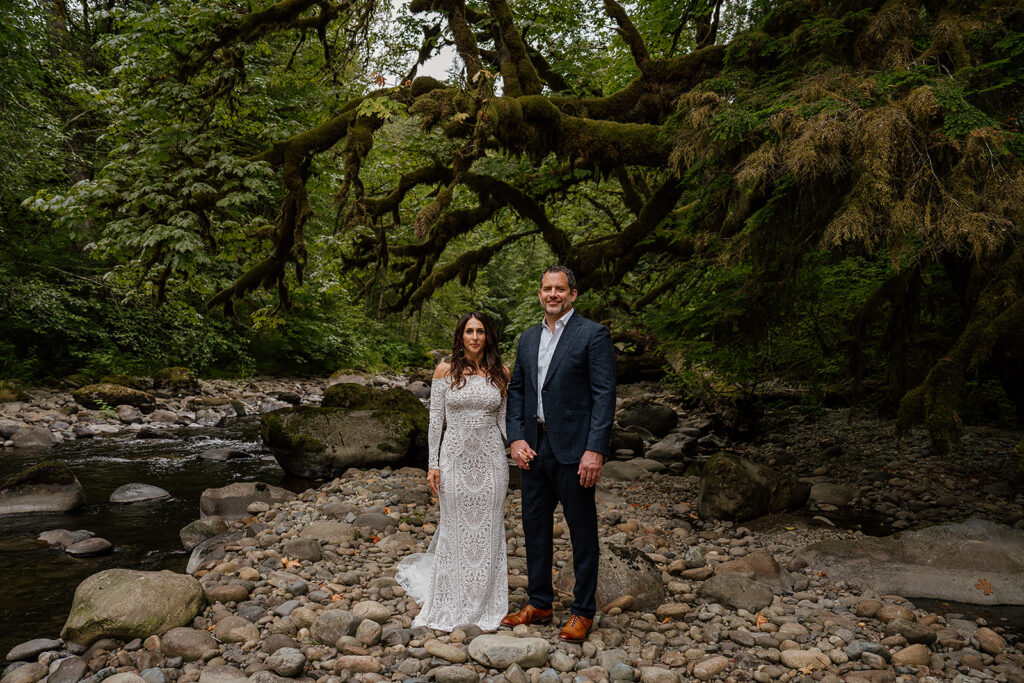 Couple stands and poses near a river bend in a mossy forest in Washington