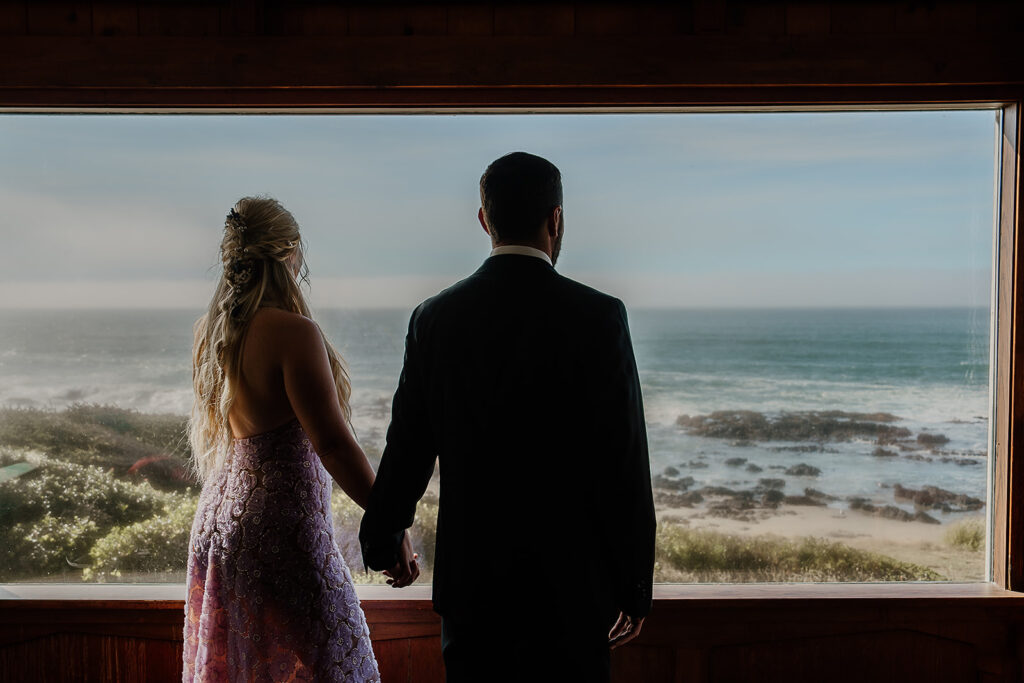 Bride and groom look out of their Oregon Airbnb to the ocean before their elopement ceremony