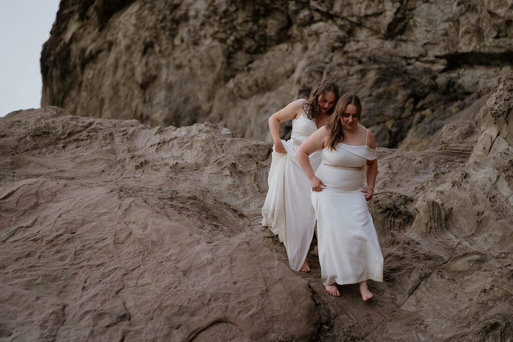 Two brides hike down a rocky cliffside to Cannon Beach