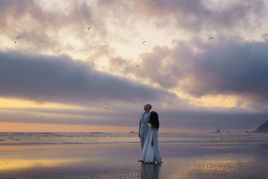 Bride and groom look up at seagulls flying while walking along Cannon Beach at sunrise