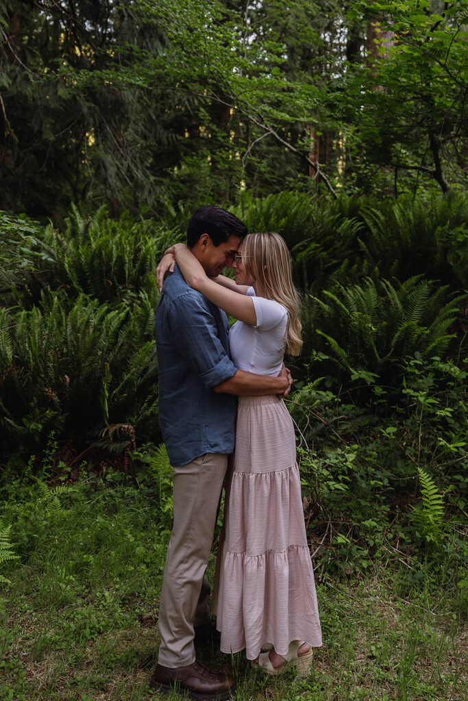 Couple stands in front of fern in a Washington forest during their lake engagement photos